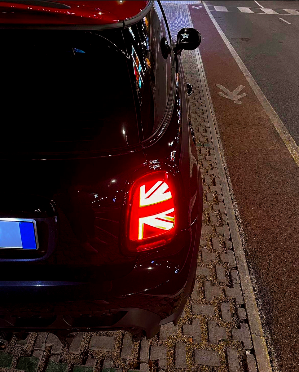 A car with illuminated Union Jack taillight is parked on a cobblestone road. It is positioned next to a bike lane symbol in a nighttime urban environment.