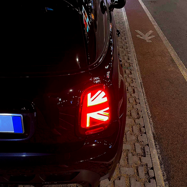 A car with illuminated Union Jack taillight is parked on a cobblestone road. It is positioned next to a bike lane symbol in a nighttime urban environment.