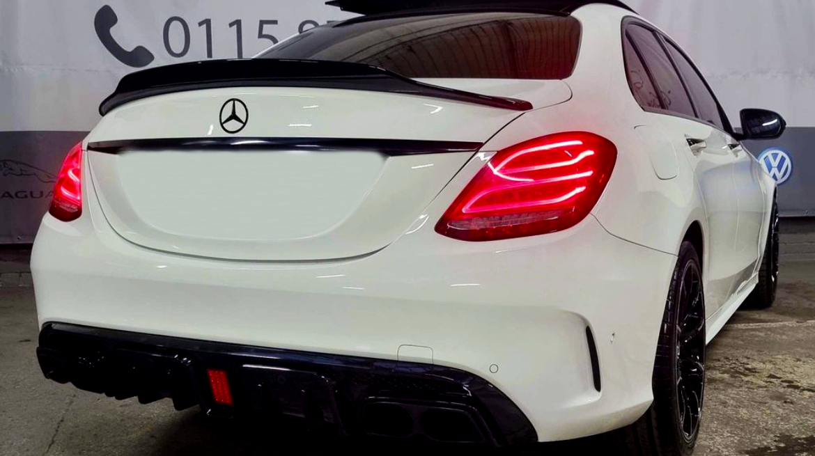 A white Mercedes-Benz car is parked inside a garage, showcasing its illuminated rear lights. The background displays a wall with phone numbers beginning with "0115" and logos, including Volkswagen and Jaguar.