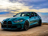 A teal sedan is parked on a sandy coastal road, with the ocean and a cloudy sky in the background, capturing a serene seaside setting.