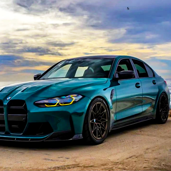 A teal sedan is parked on a sandy coastal road, with the ocean and a cloudy sky in the background, capturing a serene seaside setting.