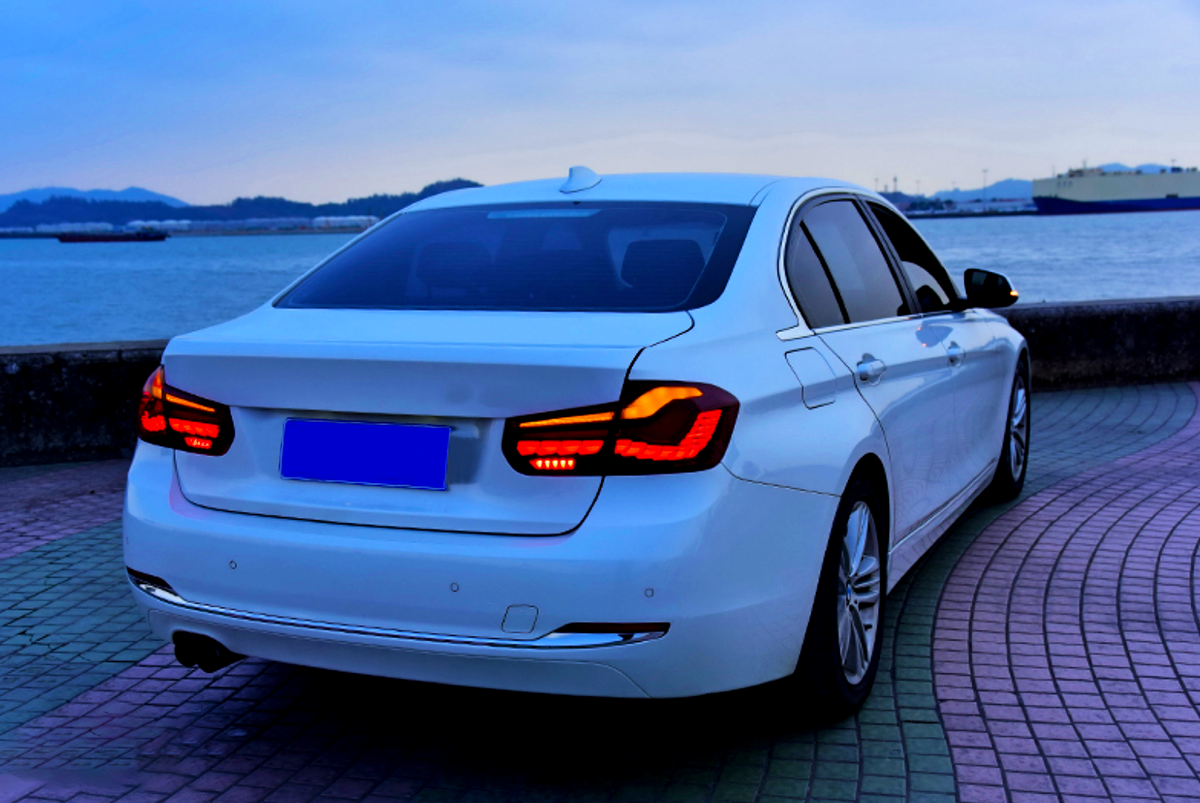 A white sedan with illuminated taillights is parked on a curved, tiled path by the waterfront. In the background, there are distant hills and a large ship on the water.