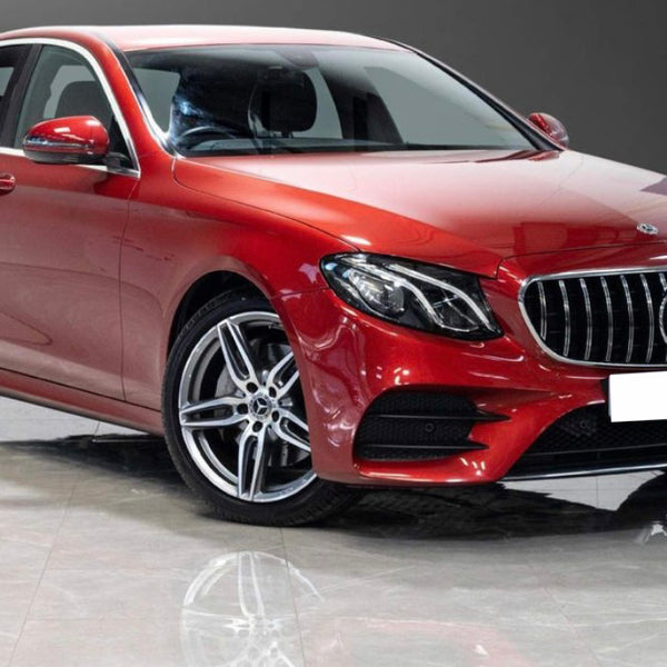 A red Mercedes-Benz sedan is parked indoors on a glossy floor, with bright lighting highlighting its sleek design and silver alloy wheels. The background is a plain dark wall.