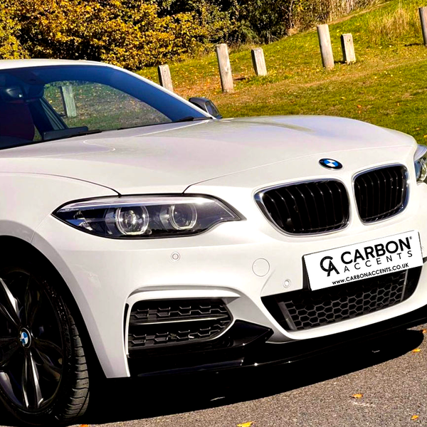 A white BMW parked on a road, angled with a green grassy background. The license plate reads “CARBON ACCENTS www.CARBONACCENTS.co.uk.” The sun casts shadows, highlighting the car’s glossy finish.