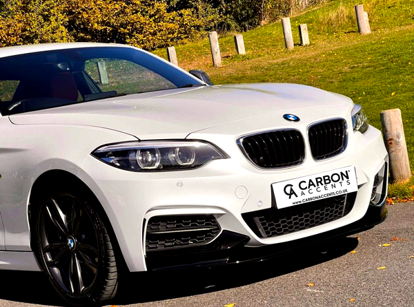A white BMW parked on a road, angled with a green grassy background. The license plate reads “CARBON ACCENTS www.CARBONACCENTS.co.uk.” The sun casts shadows, highlighting the car’s glossy finish.