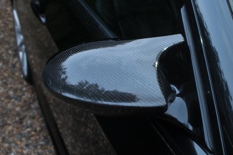 A carbon fiber car side mirror reflects its textured surface, attached to a sleek, black vehicle, with a gravel driveway visible nearby.