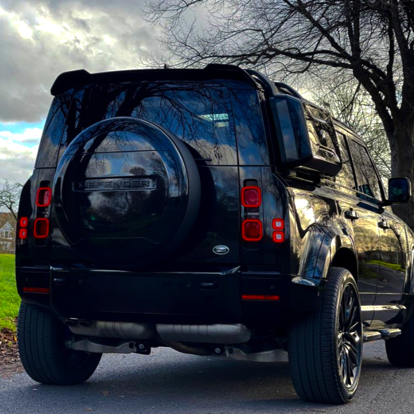 A black SUV is parked on a road, with "DEFENDER" on the rear tire cover. Trees without leaves line the path under a cloudy sky.