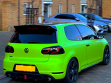 A bright green Volkswagen GTI is parked on a cobblestone street near tan brick houses, with blacked-out windows and a rear spoiler. Other cars are parked nearby.