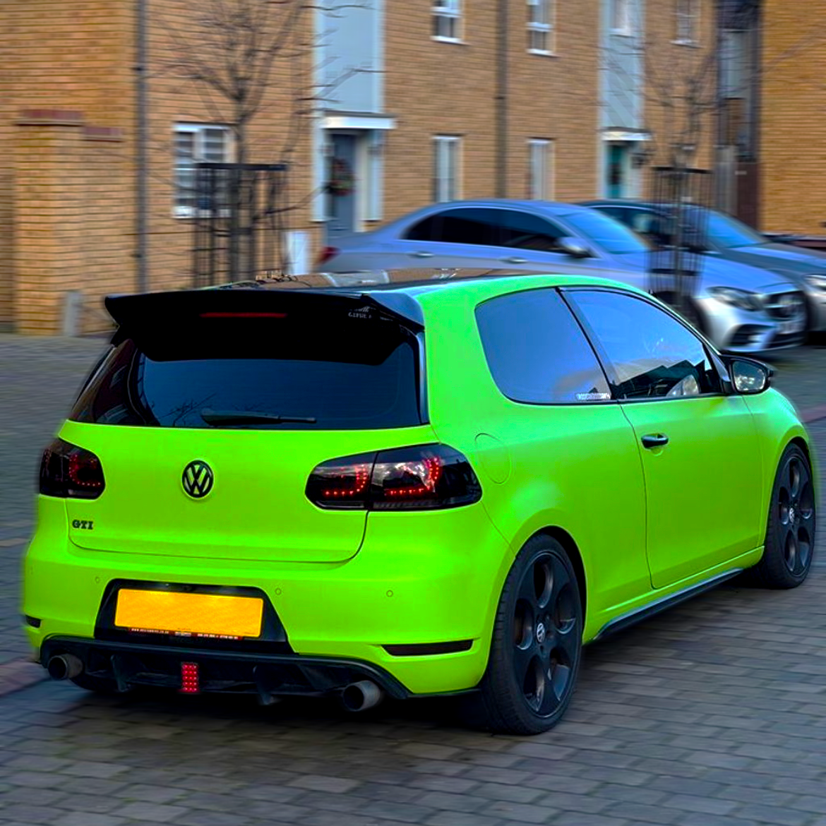 A bright green Volkswagen GTI is parked on a cobblestone street near tan brick houses, with blacked-out windows and a rear spoiler. Other cars are parked nearby.