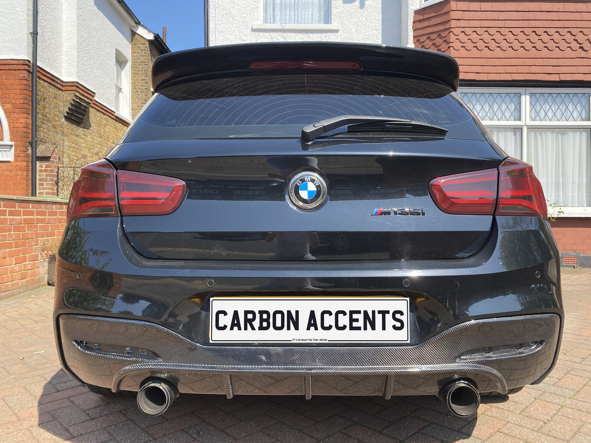 A black BMW M135i is parked on a driveway, featuring dual exhausts and a "CARBON ACCENTS" license plate. Surrounding houses display brick and white exteriors under a clear blue sky.