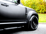 A black vehicle parked on a road features sleek body panels and large, textured tires. Trees with yellow-green foliage provide a natural backdrop.