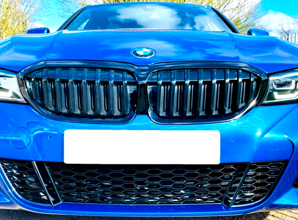 A blue car's front view showcases its black grille and logo, under a partly cloudy sky. License plate is blank, surrounded by trees and road surface visible below.