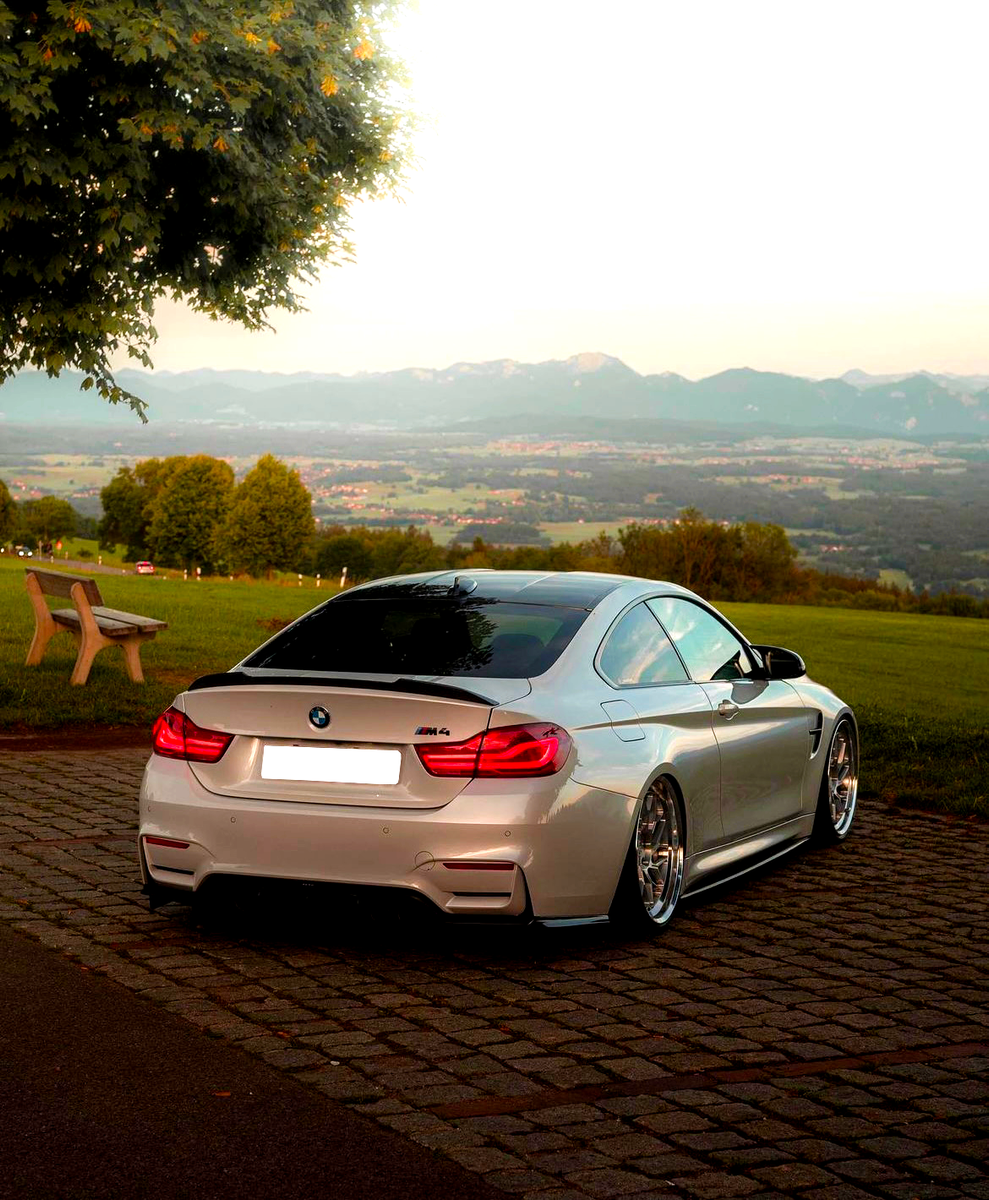 A silver BMW M4 is parked on cobblestones, surrounded by green fields and distant mountains under a clear sky with a tree and bench nearby.