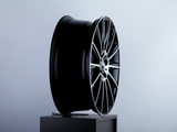 A sleek, black car wheel rim with intricate silver spokes sits upright on a gray pedestal in a minimalistic, light blue-gray setting.