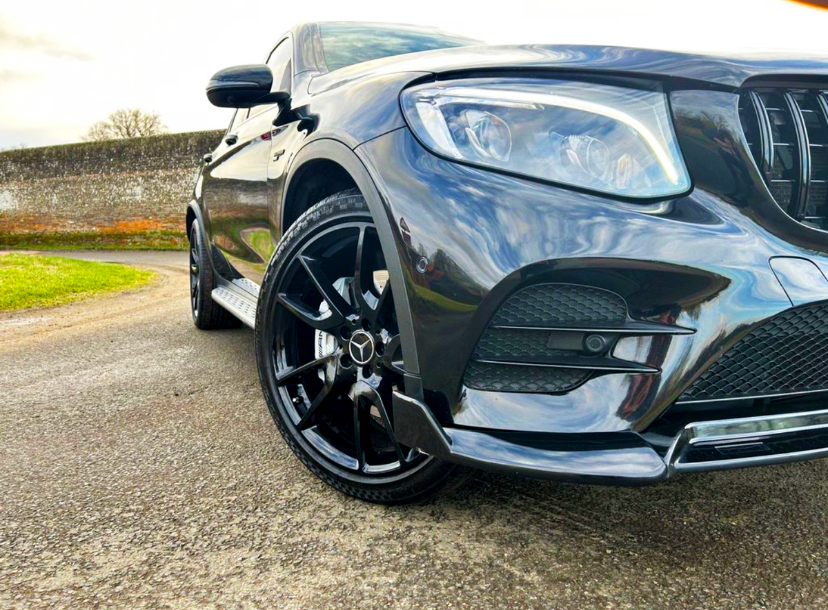 A sleek black car is parked on a paved area, featuring shiny alloy wheels and a modern headlamp design, with a stone wall and green grass in the background.