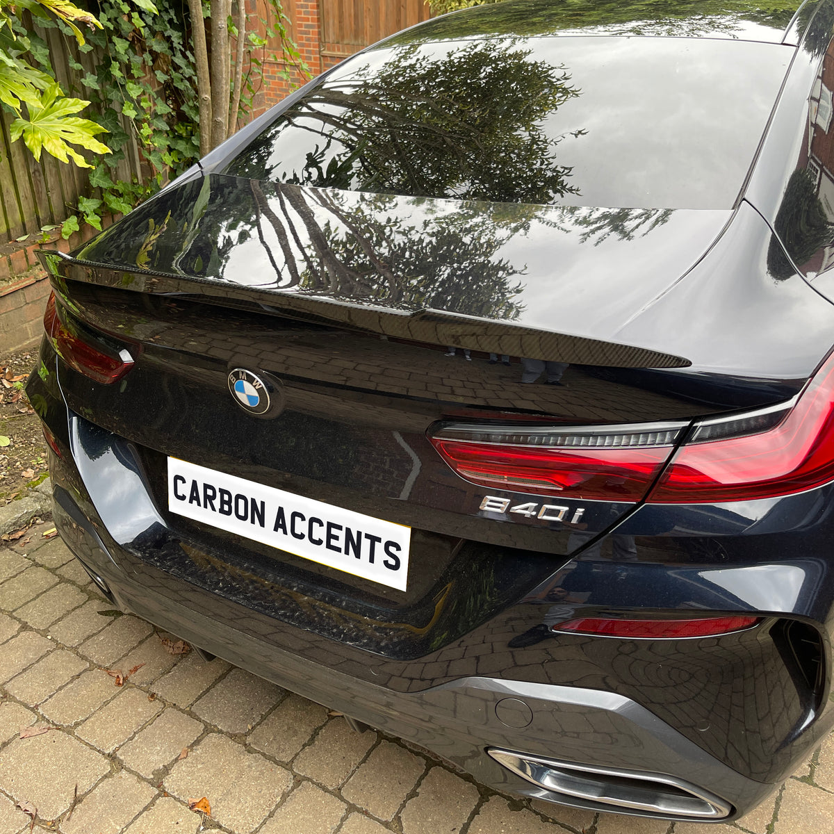 A black BMW 840i is parked on a stone-tiled driveway near a wooden fence and bushes. The license plate reads "CARBON ACCENTS." Trees reflect on the car's gloss.