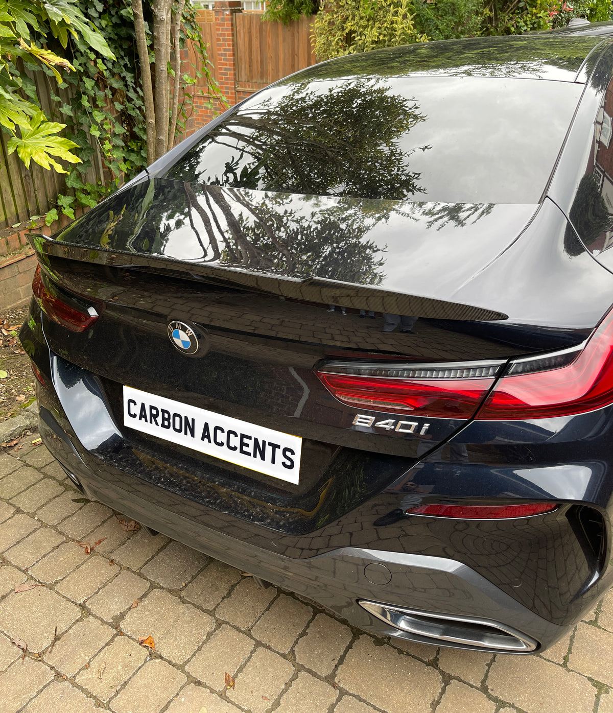A black BMW 840i is parked on a stone-tiled driveway near a wooden fence and bushes. The license plate reads "CARBON ACCENTS." Trees reflect on the car's gloss.