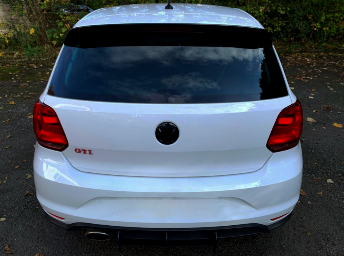 A white car is parked on an asphalt surface surrounded by fallen leaves. The trunk displays the text "GTI" in red, with rear lights and a visible exhaust pipe.