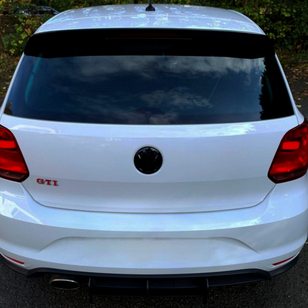 A white car is parked on an asphalt surface surrounded by fallen leaves. The trunk displays the text "GTI" in red, with rear lights and a visible exhaust pipe.