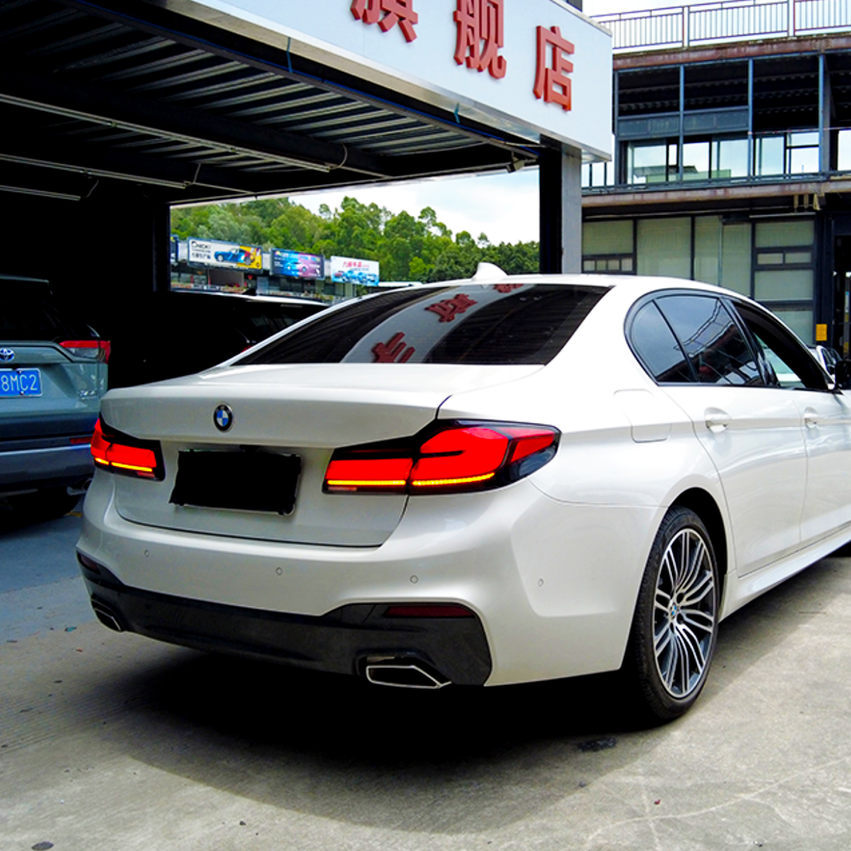 A white BMW sedan is parked outside a building with a sign in Chinese characters. Several other vehicles are nearby in a parking area, including a gray SUV.