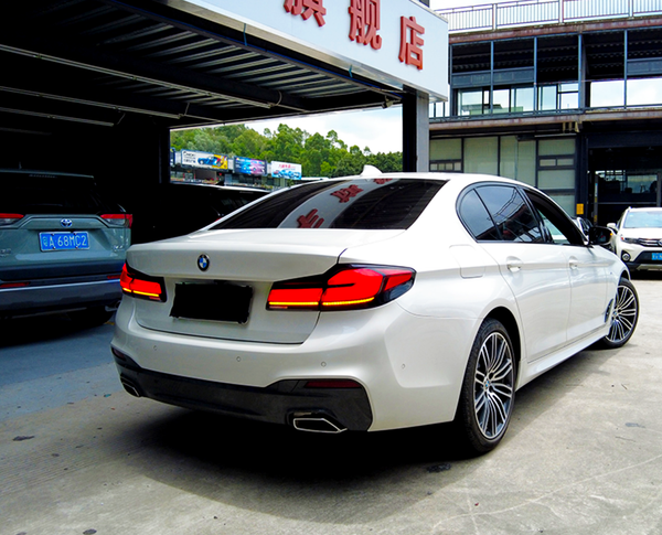 A white BMW sedan is parked outside a building with a sign in Chinese characters. Several other vehicles are nearby in a parking area, including a gray SUV.