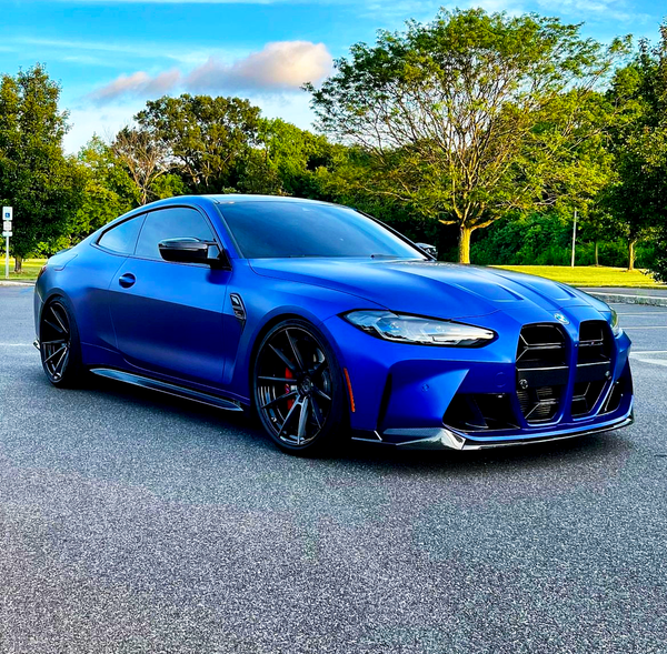 A sleek blue sports car is parked on a road, surrounded by lush green trees under a clear blue sky.