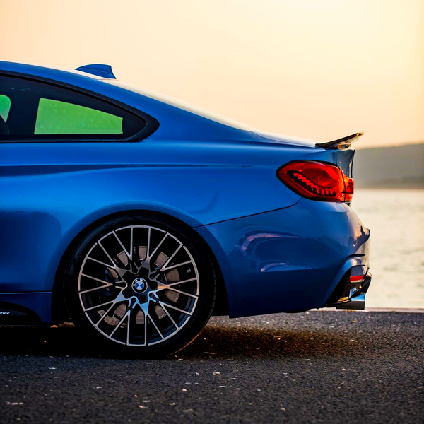 A blue sports car parked by a waterfront, showcasing its rear section with distinctive taillights and a prominent wheel in the foreground, against a serene sunset backdrop.