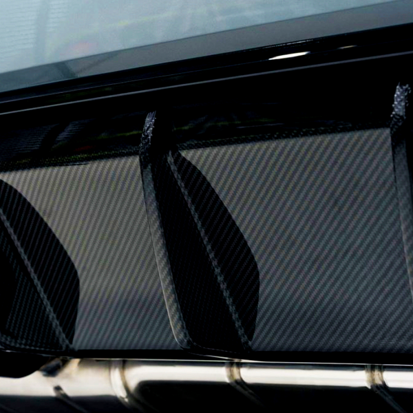 Carbon fiber rear diffuser with three vertical fins, flanked by metallic exhaust pipes, is mounted under a car's bumper in a well-lit garage setting.