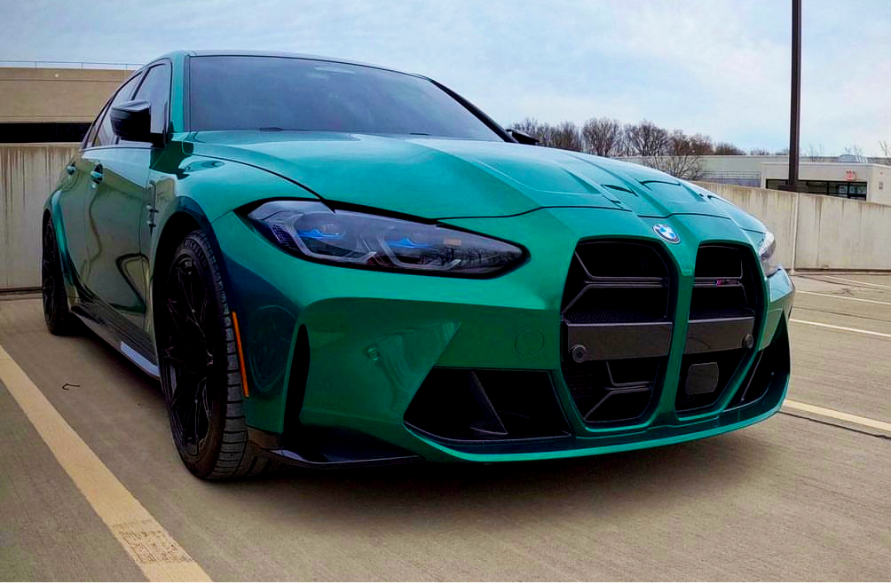 A green BMW car is parked on a rooftop parking deck, with a clear sky and bare trees in the background.