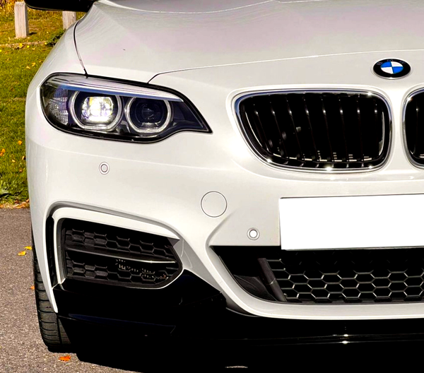 A white car's front showcases modern headlights and a signature grille. It's parked on asphalt, with grass and fallen leaves visible in the background.