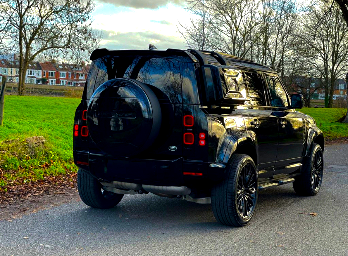 A black SUV is parked on a suburban street, featuring a rear-mounted spare tire; residential buildings and bare trees appear in the background under a cloudy sky.