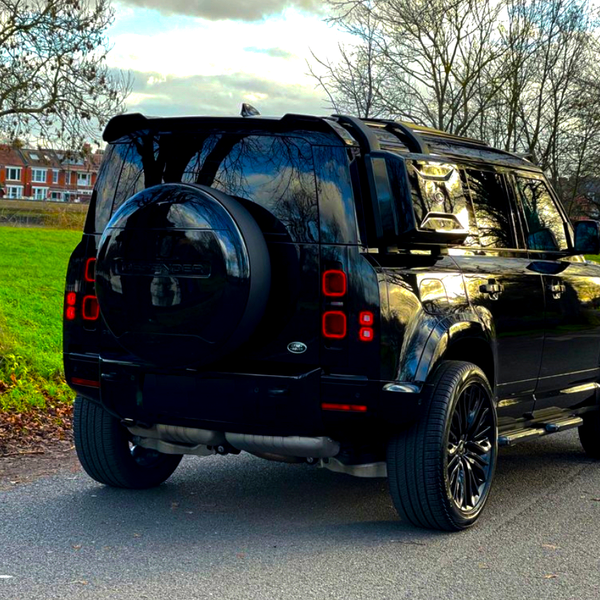 A black SUV is parked on a suburban street, featuring a rear-mounted spare tire; residential buildings and bare trees appear in the background under a cloudy sky.