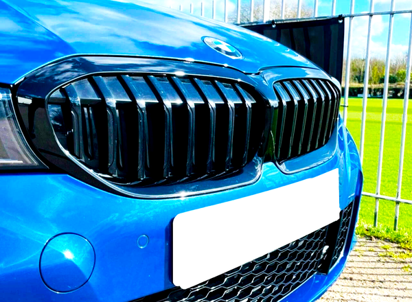 A blue car's front grille is shown, featuring a prominent emblem, parked near a metal fence with a grassy field in the background.
