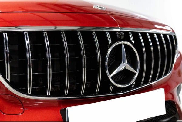A chrome-accented grille with a prominent Mercedes-Benz emblem is centered on a red car's front, highlighted in a well-lit environment. It shows sleek vertical lines and a blank license plate.