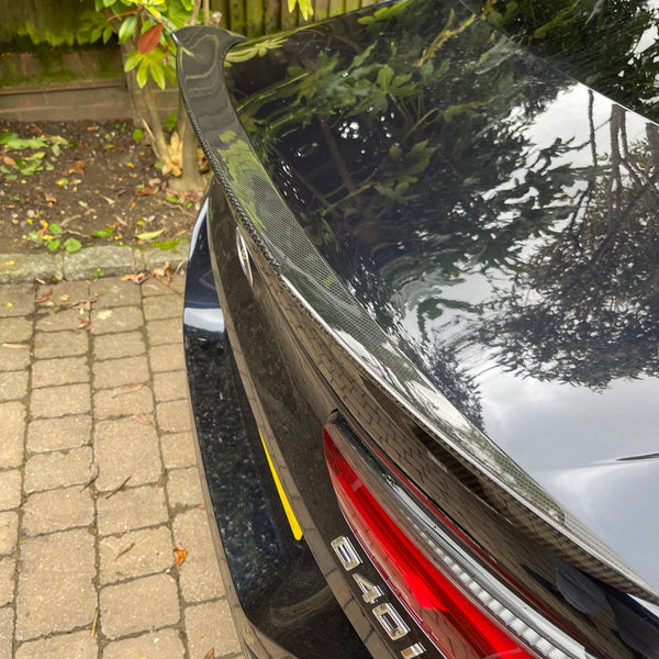 A car with a carbon fiber spoiler reflects foliage above, showing badge "640i" beside red tail lights, parked on a cobblestone driveway with a wooden fence and leaves nearby.