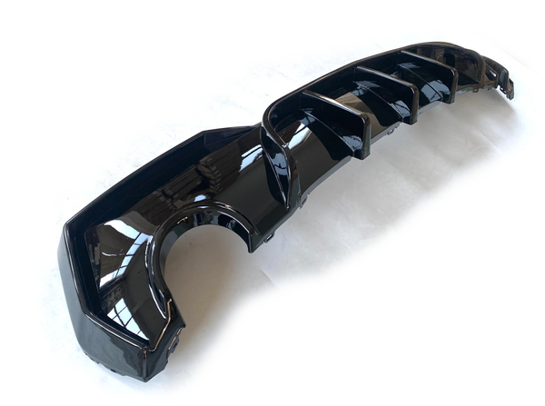 A glossy black car bumper diffuser with aerodynamic fins is lying on a white surface, reflecting light from above in a well-lit room.