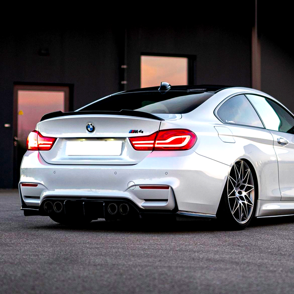 A sleek white BMW M4 with illuminated taillights sits stationary on a smooth asphalt surface, surrounded by a dimly lit industrial backdrop, featuring a partially visible building.