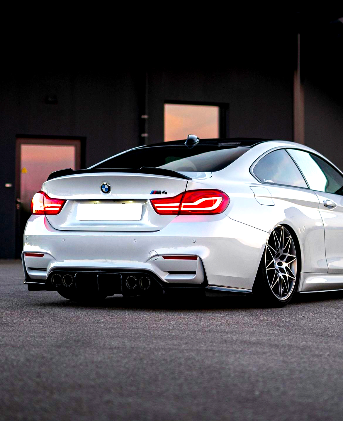A sleek white BMW M4 with illuminated taillights sits stationary on a smooth asphalt surface, surrounded by a dimly lit industrial backdrop, featuring a partially visible building.