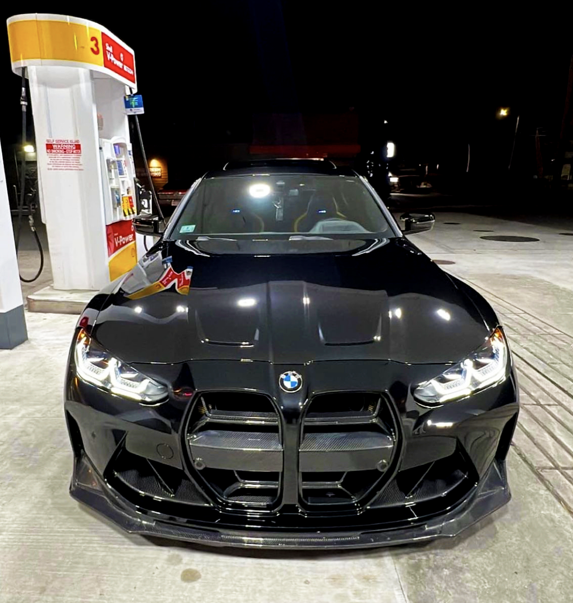 A black car, parked at a gas station at night, faces forward. It is under a Shell gas pump with visible signage that reads "Self Service Lane" and "V-Power."