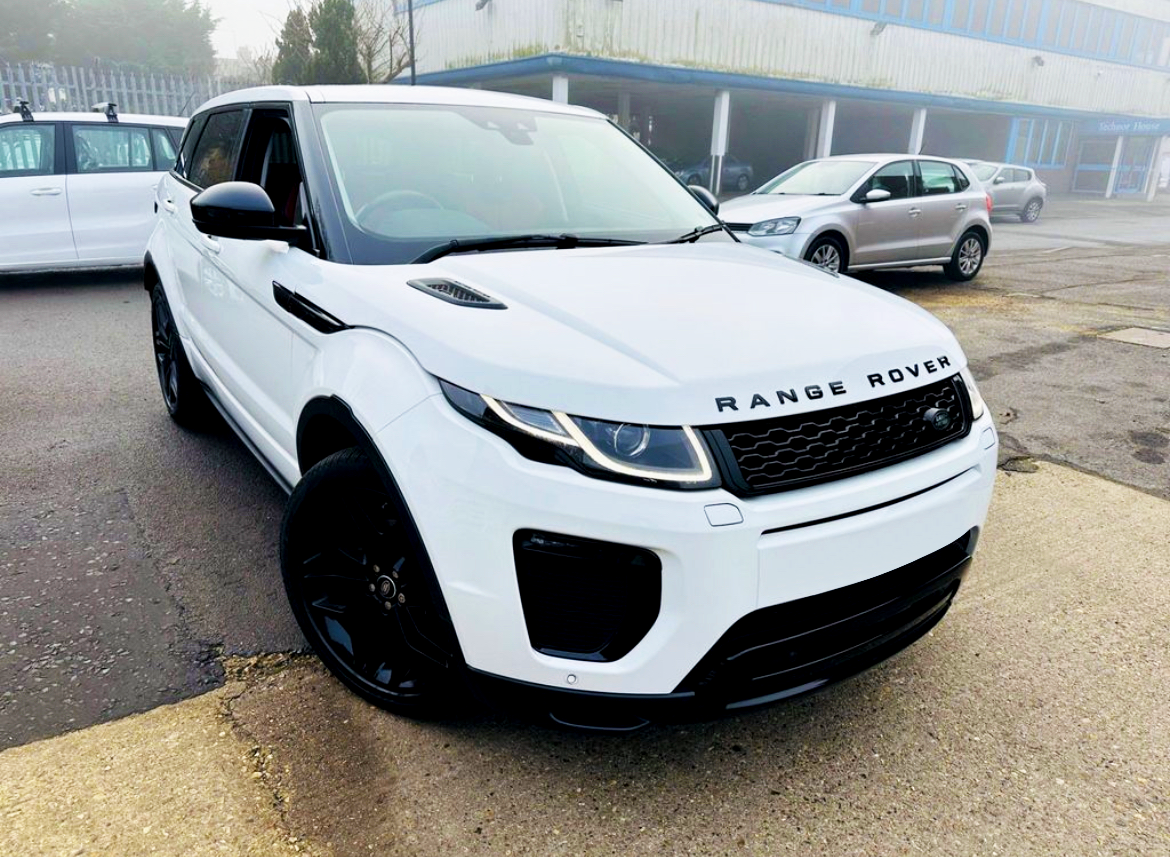 A white Range Rover parked on an asphalt surface near other vehicles in an industrial setting, with misty weather in the background. The text reads "RANGE ROVER" on the front.