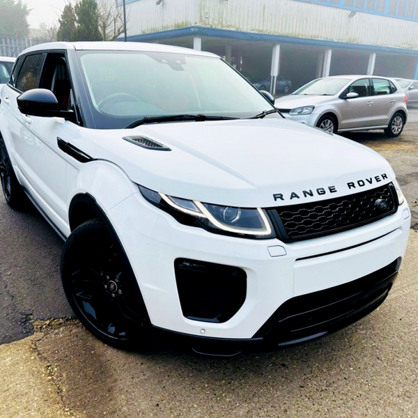 A white Range Rover parked on an asphalt surface near other vehicles in an industrial setting, with misty weather in the background. The text reads "RANGE ROVER" on the front.