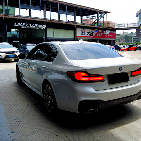 A silver BMW sedan is parked in a bustling urban setting, surrounded by other cars near buildings. Signs read "LIKE CLUB," "MO CLUB," and more in the background.