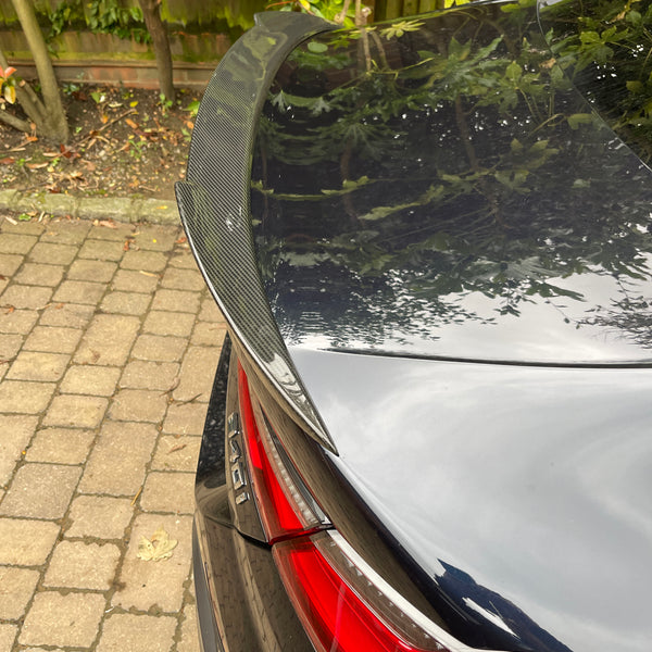 A black car trunk with a carbon fiber spoiler is parked on a stone driveway. Red tail light is visible; trees and a wooden fence are in the background.