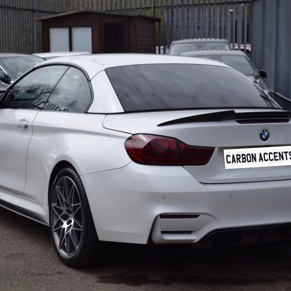 A white BMW M4 is parked in a lot beside other cars, with a license plate reading "CARBON ACCENTS." Fenced surroundings and a small structure are visible in the background.