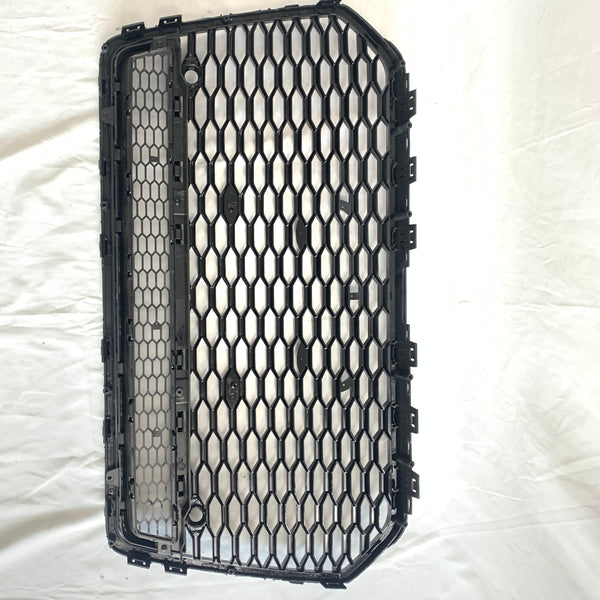 The black, hexagonal-patterned car grille is lying flat, showing its structured grid design on a white fabric backdrop, emphasizing its rectangular shape and multiple attachment clips.