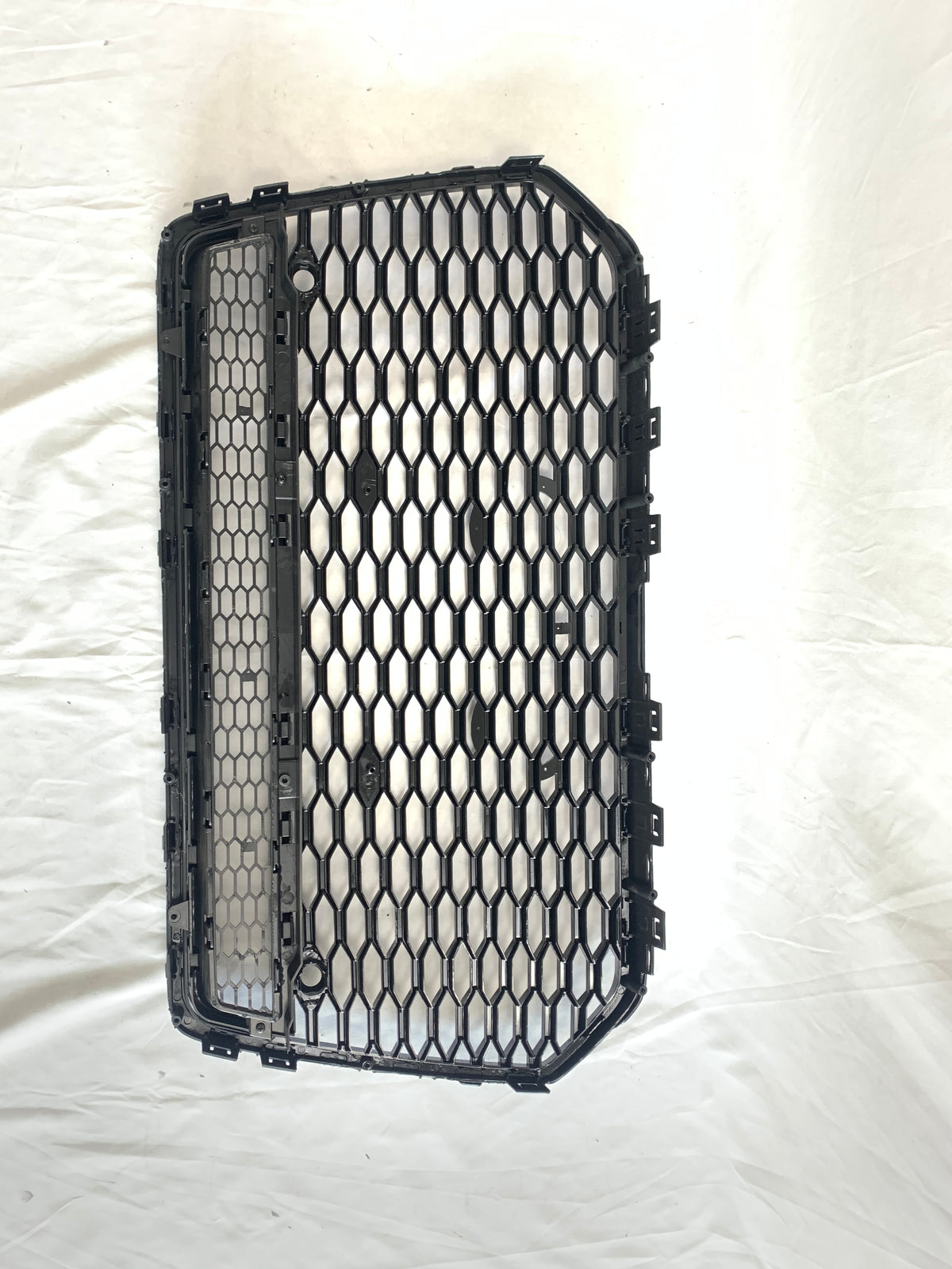 The black, hexagonal-patterned car grille is lying flat, showing its structured grid design on a white fabric backdrop, emphasizing its rectangular shape and multiple attachment clips.