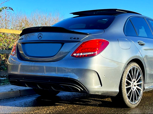 A silver Mercedes-Benz C43 car is parked on a wet road. The sunlight highlights its sleek design and red taillights, with trees and blue sky in the background.