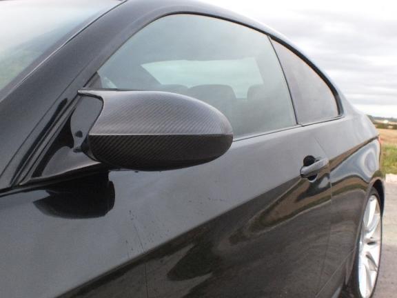A black car with a carbon fiber side mirror is parked on a road. The background features a cloudy sky and open land, creating a spacious and serene environment.