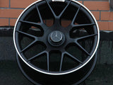 A black alloy wheel with a silver trim featuring a Mercedes-Benz logo rests against a brick wall. The AMG insignia is visible on the top part of the rim.