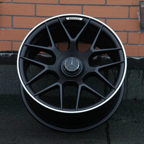 A black alloy wheel with a silver trim featuring a Mercedes-Benz logo rests against a brick wall. The AMG insignia is visible on the top part of the rim.
